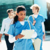 Female healthcare student in blue scrubs holds a clipboard as she walks outside with teammates and a modern glass building in the background.
