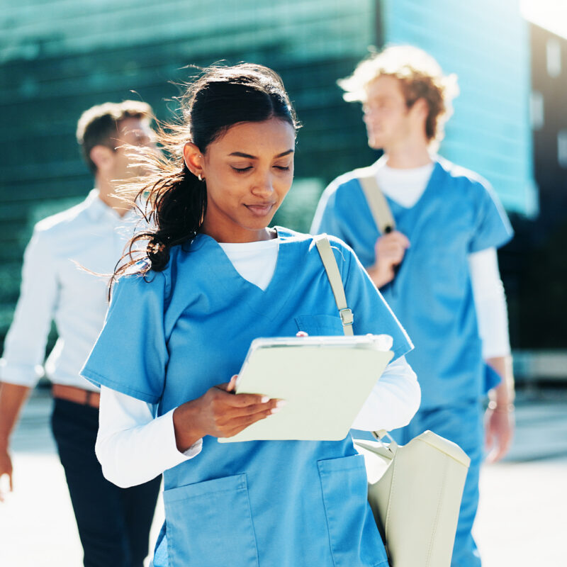 Female healthcare student in blue scrubs holds a clipboard as she walks outside with teammates and a modern glass building in the background.