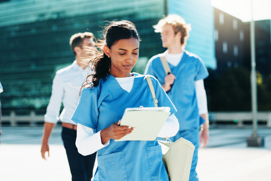 Female healthcare student in blue scrubs holds a clipboard as she walks outside with teammates and a modern glass building in the background.