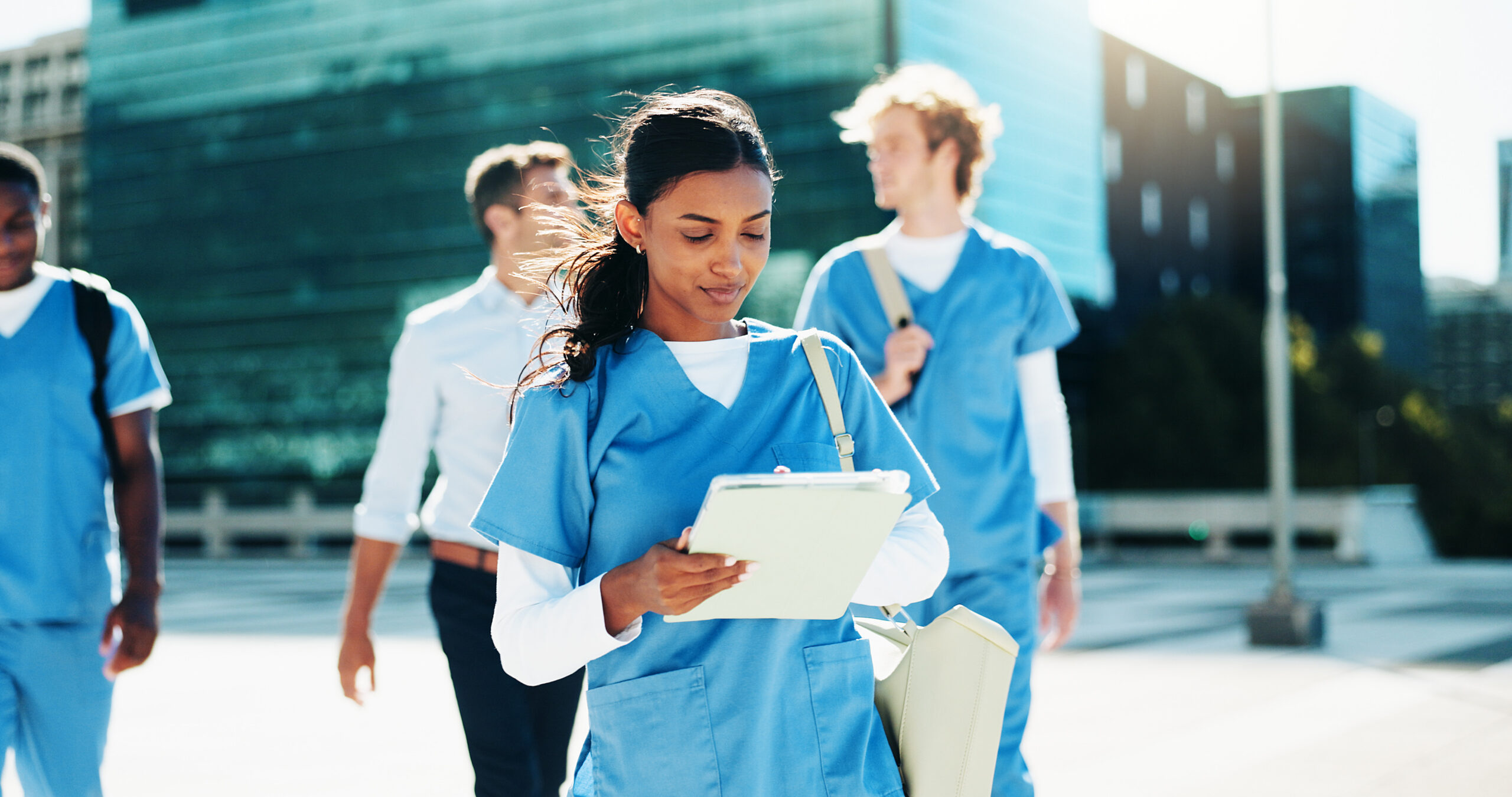Female healthcare student in blue scrubs holds a clipboard as she walks outside with teammates and a modern glass building in the background.
