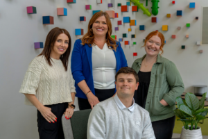Four colleagues posing for a photo in a bright office with a wall of colorful cube decorations behind them.
