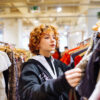 portrait of red-haired attractive young woman trying on clothes in store | The Green on Campus Drive Woman with curly red hair browsing clothing on a rack in a bright shop near shelves of garments in a store.