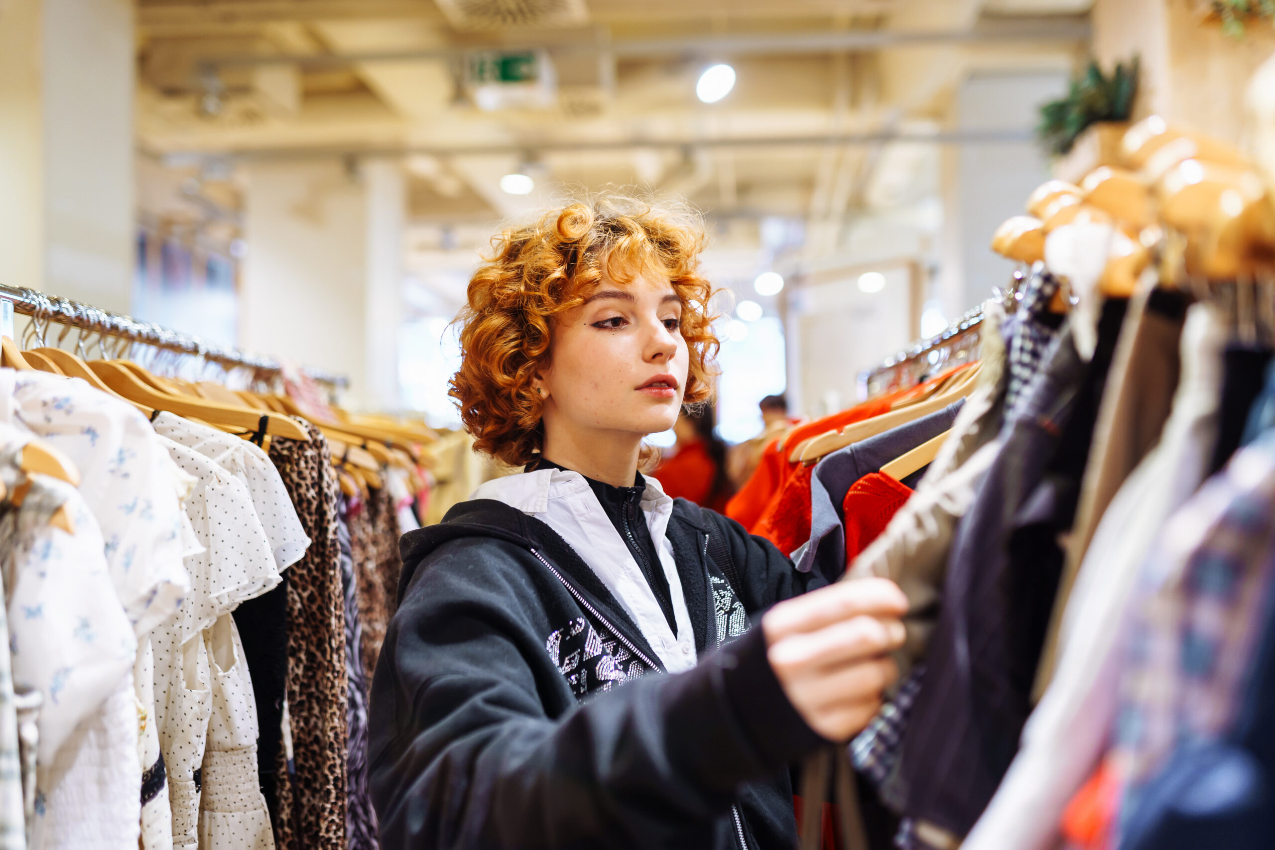 Woman with curly red hair browsing clothing on a rack in a bright shop near shelves of garments in a store.
