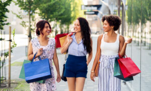 Three women smiling and chatting while carrying colorful shopping bags outdoors on a sunny day