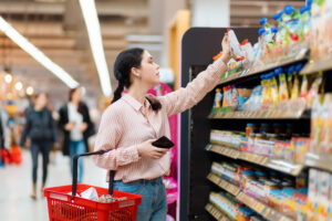 Woman reaching for a snack from a grocery store shelf while pushing a red shopping basket on the cart handle, in a bright supermarket aisle.