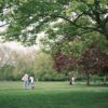 Parks Near UVU in Orem UT | The Green on Campus Drive Two people walk a small dog across a grassy park lawn under large shade trees, with others relaxing on the grass in the distance.